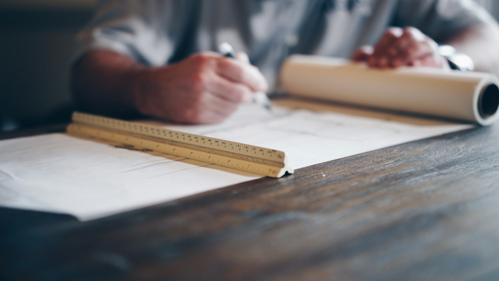 Manos dibujando un plano arquitectónico sobre un escritorio, con regla y rollo de papel